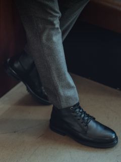 Persona con pantaloni grigi e stivali stringati in pelle nera, una gamba accavallata sull'altra, in piedi in un interno su un pavimento chiaro.