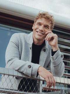 Un uomo sorridente con capelli biondi ricci si appoggia con disinvoltura a una recinzione metallica, indossa un cappotto grigio chiaro sopra una camicia nera e ha un edificio moderno sullo sfondo.