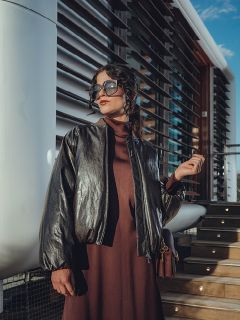 Una donna con grandi occhiali da sole e una giacca di pelle scura è appoggiata a un muro, con le mani incrociate dietro la testa, e posa con sicurezza alla luce del sole.