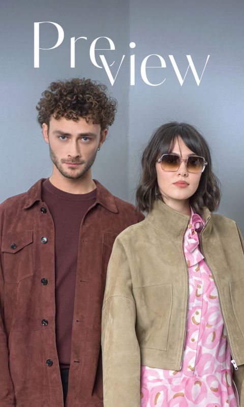 Un uomo con una giacca marrone e una donna con occhiali da sole e una giacca marrone chiaro sopra un abito rosa fantasia sono in piedi uno accanto all'altra davanti a un semplice sfondo chiaro.