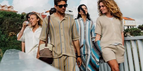 Un gruppo di cinque persone si fa un selfie all'aperto vicino all'acqua, con alberi e un tramonto sullo sfondo. Sorridono e posano in modo giocoso, vestiti in abiti casual. Sopra di loro si vedono il testo "KnowledgeCotton Apparel" e un logo a forma di gufo.