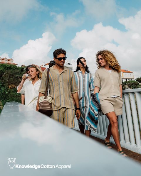 Un gruppo di cinque persone si fa un selfie all'aperto vicino all'acqua, con alberi e un tramonto sullo sfondo. Sorridono e posano in modo giocoso, vestiti in abiti casual. Sopra di loro si vedono il testo "KnowledgeCotton Apparel" e un logo a forma di gufo.
