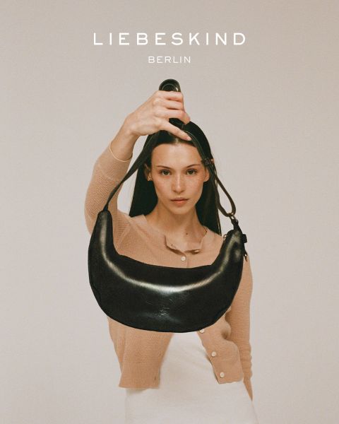 Una donna sorridente con una camicia nera a maniche lunghe e jeans indossa una borsa marrone chiaro a tracolla. Il testo "Liebeskind Berlin" appare nella parte inferiore dell'immagine.