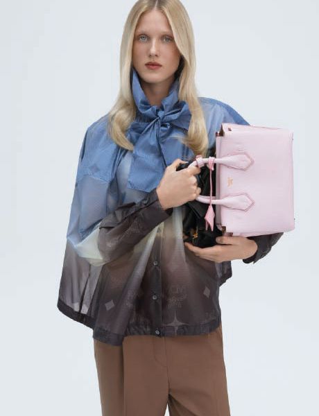 Una borsa verde di design con un discreto motivo monogrammato è appoggiata su una superficie grigia, con un oggetto geometrico blu in primo piano e un'ombra proiettata sullo sfondo.