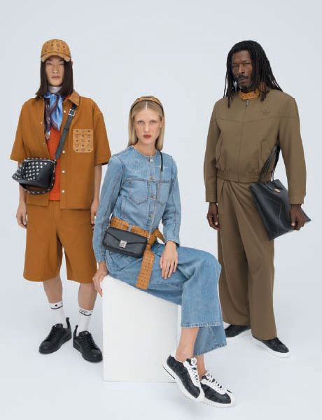 Una borsa firmata MCM blu con motivo logo bianco è appoggiata su gradini di legno decorati con colorati portachiavi a forma di animali e oggetti, tra cui un razzo, un coniglio e un orso.