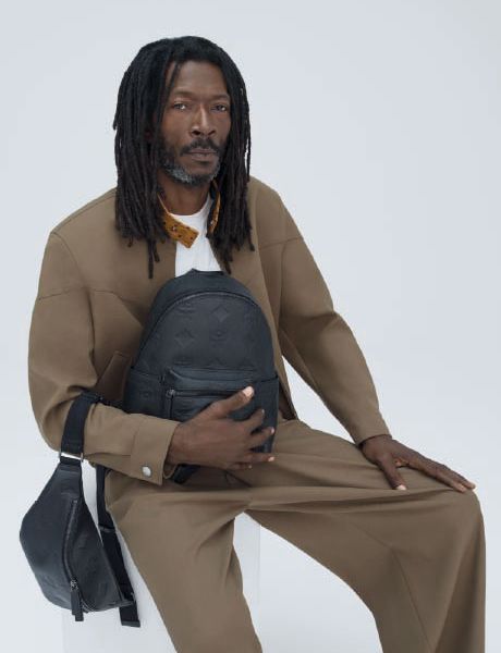 Uno zaino di design verde e una borsa a tracolla abbinata sono appoggiati su una pila di scatole arancioni e marroni davanti a una parete strutturata beige e un pavimento piastrellato.