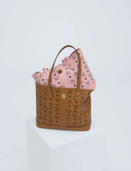 Una donna con una giacca shearling marrone chiaro, pantaloncini neri e stivali è in piedi su un gradino di pietra e tiene in mano una grande borsa. Sullo sfondo, un uomo vestito con abiti colorati sembra fluttuare orizzontalmente sopra i gradini. Dietro di loro si vedono degli alberi.