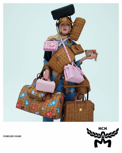 Tre donne con cappotti marrone chiaro e cappelli arancioni camminano per una strada, portando borse MCM blu con un logo MCM bianco in primo piano. L'immagine ha un effetto specchio che fa apparire la scena duplicata.