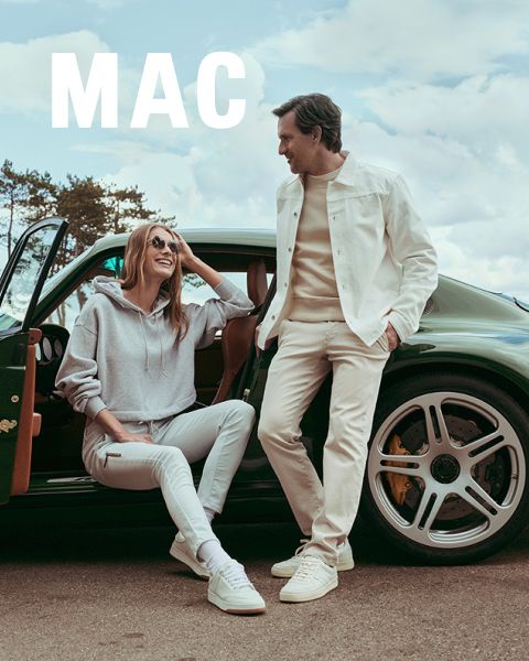 Una donna con una giacca di pelliccia e pantaloni ampi è in piedi accanto a un uomo con una giacca beige e pantaloni color kaki. Entrambi posano con sicurezza all'aperto di fronte a una ringhiera di pietra sotto un cielo blu. La parola "MAC" appare in basso.