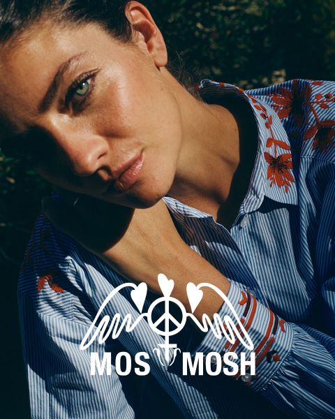 Una donna con lunghi capelli biondi ondulati indossa una camicia verde con motivi bianchi. Guarda sopra la sua spalla e si trova di fronte a un edificio moderno. Il logo "MOS MOSH" si trova nell'angolo in alto a sinistra.