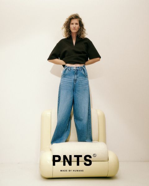 Una donna è in piedi con sicurezza davanti a una porta di vetro, indossa una giacca beige, un top nero, jeans larghi blu e scarpe beige. Il testo "PNTS MADE BY HUMANS" è visualizzato nella parte inferiore dell'immagine.