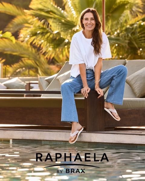 Una donna con lunghi capelli biondi, che indossa una camicia grigia a maniche lunghe e jeans scuri, è in piedi sorridendo a una ringhiera con uno sfondo azzurro. Accanto a lei appare il testo "EUREX di BRAX".