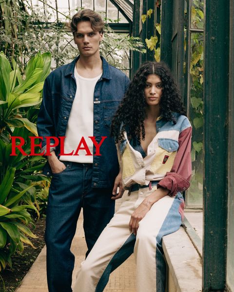 Un uomo e una donna siedono su una scala di legno in un interno, vestiti con abiti scuri ed eleganti. La parola "REPLAY" è sovraimpressa in modo prominente al centro dell'immagine in lettere maiuscole rosse.