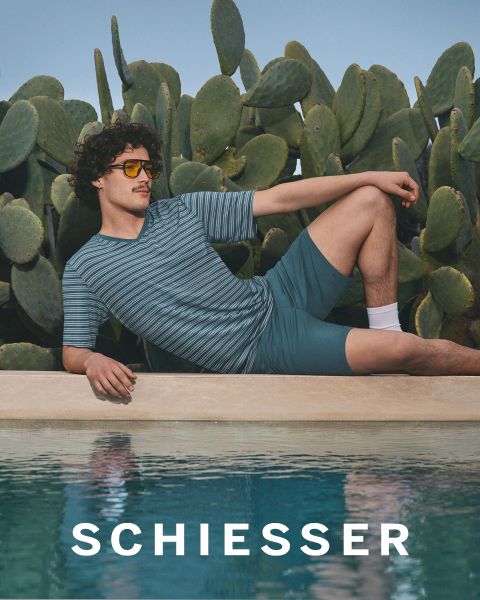 Una donna e un uomo in abbigliamento da casa grigio chiaro siedono vicini su un tappeto e sorridono dolcemente alla fotocamera. Il marchio "SCHIESSER" è visibile nella parte inferiore dell'immagine.