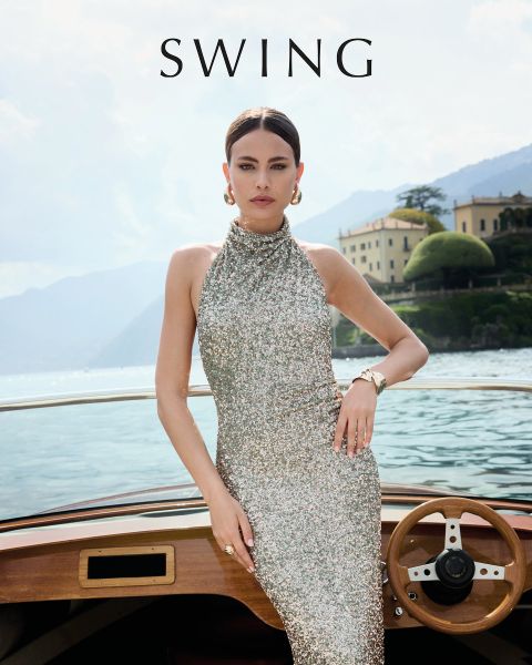 Una donna in un abito di seta color verde salvia è seduta su una sedia color crema davanti a uno sfondo floreale. Indossa tacchi dorati, orecchini vistosi e un braccialetto. La parola "SWING" appare nella parte inferiore dell'immagine.