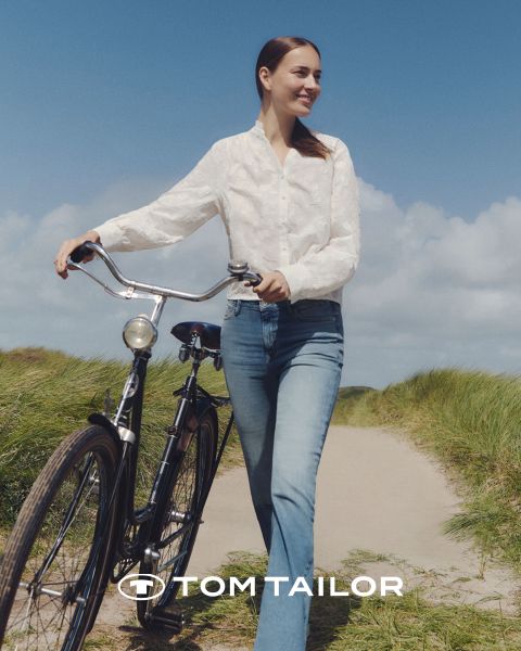 Una donna sorridente con un maglione color castagna e un uomo con un maglione blu navy sono in piedi fianco a fianco su una spiaggia. Entrambi indossano jeans. Il cielo è limpido e blu. Il logo "Tom Tailor" appare in basso.