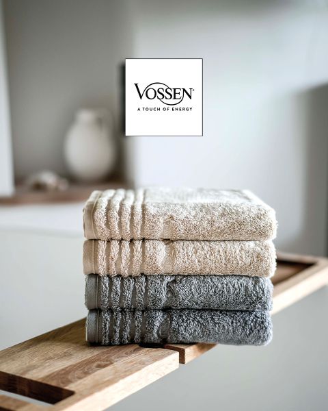 Una donna con i capelli biondi corti, avvolta in un asciugamano grigio, è seduta su un divano chiaro e guarda la fotocamera. Nell'angolo inferiore sinistro si vede il logo Vossen.