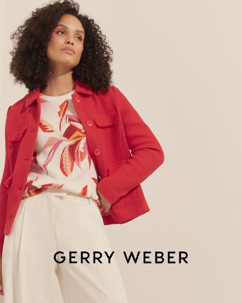 Una donna con i capelli ricci indossa una giacca rossa strutturata sopra un top floreale colorato e pantaloni color crema. Lo sfondo è semplice e in basso si vede il testo "Gerry Weber".
