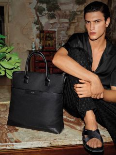 Una persona con capelli scuri è seduta su una superficie di marmo, indossa una camicia scura abbottonata e pantaloni. È a piedi nudi, con un piede accavallato sull'altro e indossa sandali neri. Accanto a lei c'è una grande borsa di pelle nera. Sullo sfondo si vedono un muro e mobili in stile vintage.