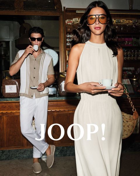Uomo e donna in abiti estivi alla moda in un caffè. L'uomo indossa una camicia beige e tiene in mano una tazza, mentre la donna con un vestito chiaro tiene un piattino. Entrambi indossano occhiali da sole. "JOOP!" è ben visibile.