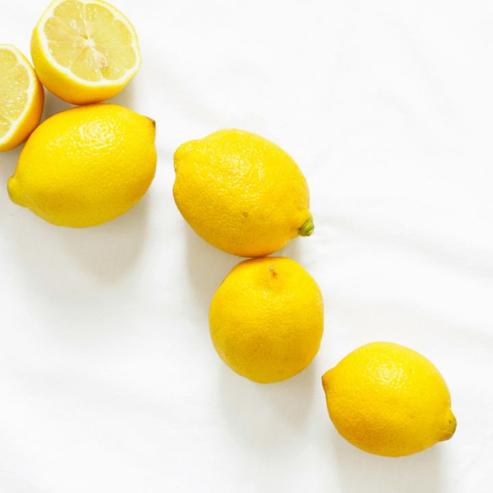 Su una superficie bianca sono disposte quattro limoni interi e due metà di limone. I limoni sono di un giallo brillante e creano un contrasto con lo sfondo.