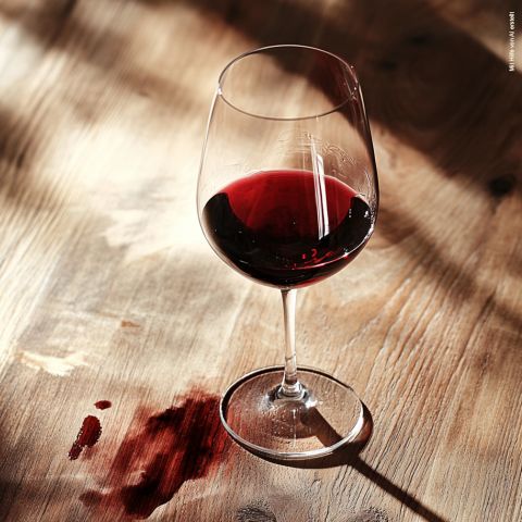 Un bicchiere di vino rosso è posato su un tavolo di legno e proietta un'ombra. Nelle vicinanze, alcune macchie di vino aggiungono un tocco di colore rosso alla superficie del legno, ulteriormente evidenziato dalla luce solare che entra.