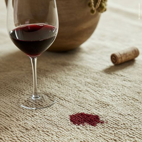 Un bicchiere di vino rosso è posato su una superficie chiara e strutturata. Nelle vicinanze si nota una macchia di vino rosso. Sullo sfondo, accanto a una ciotola di legno, un tappo di sughero giace sulla superficie.