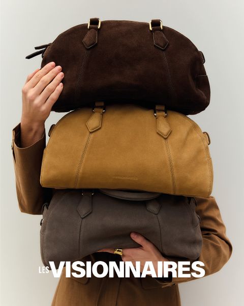 Una persona con un cappotto marrone alza una mano per coprire parzialmente il viso. Si trova di fronte a uno sfondo scuro. Nella parte superiore dell'immagine, le parole "Les Visionnaires" sono scritte in grassetto bianco.