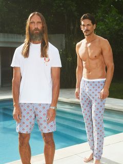 Un uomo senza camicia in un blazer nero e pantaloncini scuri è seduto su una sedia bianca su scogliere rocciose in riva al mare, con un bicchiere di vino bianco in mano, mentre le onde si infrangono sullo sfondo.