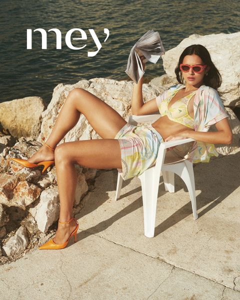 Un uomo in boxer neri e una donna in canottiera bianca e biancheria intima sono in piedi uno accanto all'altra all'aperto vicino a una ringhiera, con il mare e il cielo nuvoloso sullo sfondo. La parola "mey" è in basso.