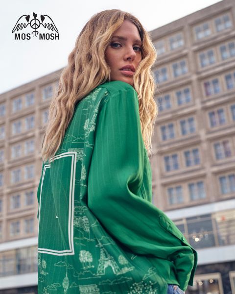 Una donna con lunghi capelli biondi ondulati indossa una camicia verde con motivi bianchi. Guarda sopra la sua spalla e si trova di fronte a un edificio moderno. Il logo "MOS MOSH" si trova nell'angolo in alto a sinistra.