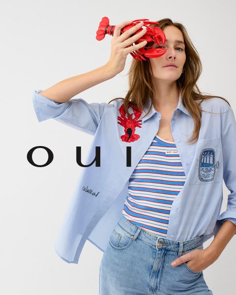 Una donna che indossa un maglione rosso con un cuore bianco, jeans blu e un cappello bianco posa davanti a un muro chiaro. Nella parte inferiore dell'immagine c'è la parola "oui" in lettere bianche.