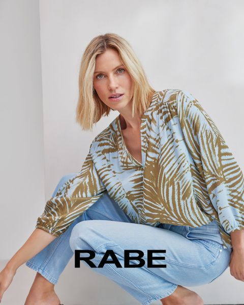 Una donna posa davanti a un muro bianco, indossa un gilet rosso senza maniche sopra una camicia a righe rosse e bianche e pantaloni scuri. Nella parte inferiore dell'immagine si legge la parola "RABE".