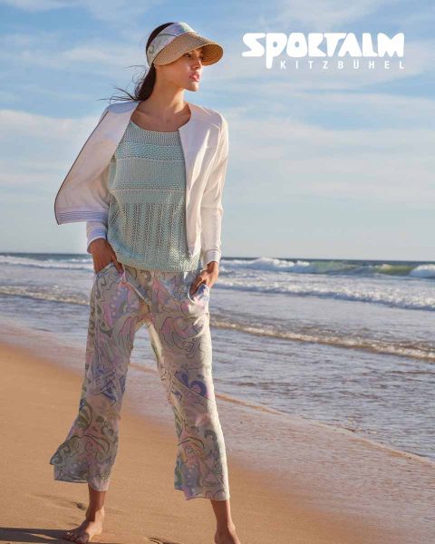 Una donna è in piedi su una spiaggia. Indossa un top in maglia azzurro, una giacca bianca e pantaloni fantasia. Porta una visiera e guarda verso il mare. Nell'angolo in alto a destra c'è scritto "SPORTALM KITZBÜHEL".