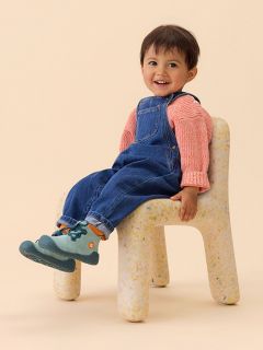 Un bambino piccolo con una camicia a righe e una salopette di jeans è seduto con una gamba all’aperto e mostra la suola blu strutturata della sua scarpa.