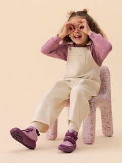 Un bambino sorridente con un cappello verde, una camicia a righe e un gilet è seduto su un blocco color pesca davanti a uno sfondo giallo, indossando stivali e pantaloni chiari.