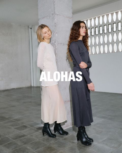 Una donna in un top bianco che lascia scoperto l'ombelico e pantaloni beige con dettagli arancioni è seduta su uno sgabello. Si trova in una stanza rustica con grandi finestre. Al centro si vede il testo "ALOHAS".