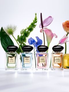 Quattro flaconi di profumo sono esposti sopra e intorno a un piedistallo beige. I flaconi sono quadrati con tappi arrotondati e sono nei colori nero e rosso. Una parete bianca ondulata e strutturata fa da sfondo.