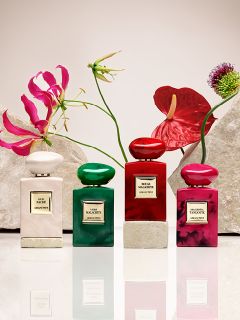 Otto flaconi di profumo in vari colori, tra cui rosso, verde, blu, rosa e nero, sono disposti su piedistalli beige davanti a uno sfondo chiaro. Ogni flacone ha un'etichetta con scritte dorate.