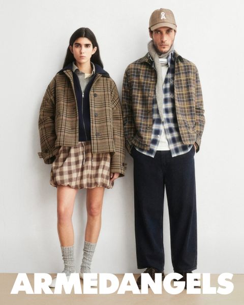 Due giovani adulti in abbigliamento di jeans stanno insieme all'aperto, uno con il braccio alzato, l'altro con una valigia coperta di adesivi. In basso, in grassetto e scritta bianca, c'è la scritta "ARMEDANGELS".