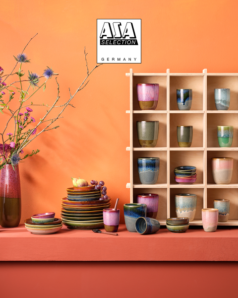 Caraffe color ambra con bicchieri abbinati, riempite con un liquido marrone, sono posizionate su un vassoio dorato davanti a uno sfondo suggestivo con texture grigia. In alto è raffigurato il logo di ASA Selection Germany.