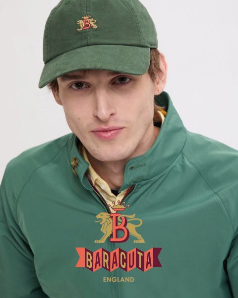 Una persona con capelli corti e scuri e un orecchino, che indossa una giacca scura sopra una camicia bianca, guarda la fotocamera. L'interno della giacca è a quadri rossi. Sul davanti è raffigurato il logo Baracuta con il leone e la "B".