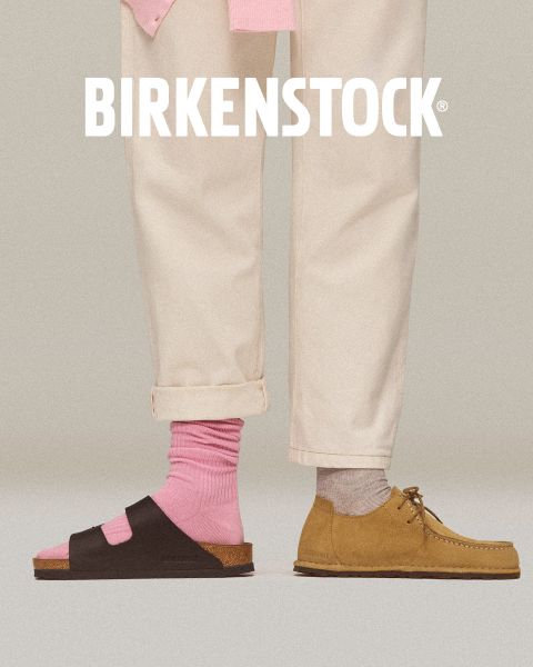Un paio di zoccoli Birkenstock beige con cinturini con fibbia su un pavimento di mattoni rossi accanto a una parete piastrellata rossa geometrica. Il logo bianco Birkenstock si trova nella parte inferiore dell'immagine.