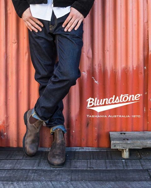 Una persona con stivali Blundstone marroni consumati si trova su una barca logora vicino a delle corde, con acqua e rocce sfocate sullo sfondo.
