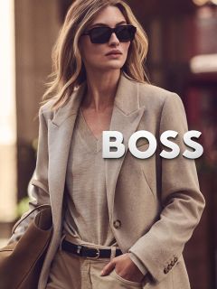 Una donna con lunghi capelli scuri e trucco neutro indossa una giacca shearling marrone e un dolcevita beige e guarda direttamente nella fotocamera con un'espressione serena, su uno sfondo semplice.