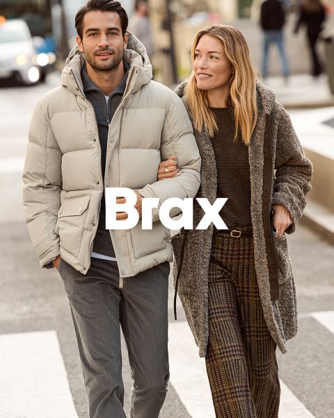 Una donna con un cappotto fantasia e un maglione rosso è in piedi accanto a un uomo con un cappotto invernale blu navy e pantaloni beige. Sono appoggiati a un muro di pietra all'aperto. La parola "Brax" appare nella parte inferiore dell'immagine.