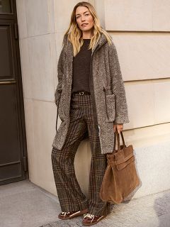 Una donna con i capelli lunghi è in piedi su un marciapiede in città. Indossa un maglione nero con strisce bianche e pantaloni neri a gamba larga e tiene in mano una grande pochette nera, mentre è illuminata dal sole da dietro.