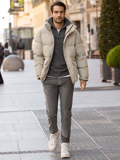 Un uomo con capelli scuri corti e la barba è appoggiato a una ringhiera di pietra all'aperto. Indossa una giacca a quadri rossi e bianchi sopra un maglione color crema e jeans blu. Sullo sfondo si vedono edifici urbani.