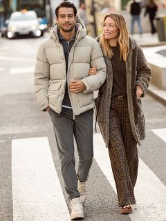 Una donna con un cappotto di pelliccia fantasia e un top rosso sorride accanto a un uomo con una giacca invernale blu navy e un maglione beige. Sono in piedi all'aperto vicino a un muro di pietra e la donna tiene in mano una borsa marrone.
