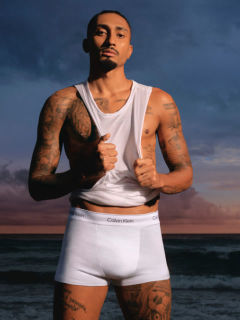 Un uomo con una giacca nera e jeans è in piedi in uno stadio e guarda di lato con le mani in tasca. Sullo sfondo si vedono i posti a sedere dello stadio e un grande edificio.